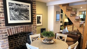 A dining area with a brick fireplace, a round table with a polka dot tablecloth, white chairs, and a basket of greenery on the table.