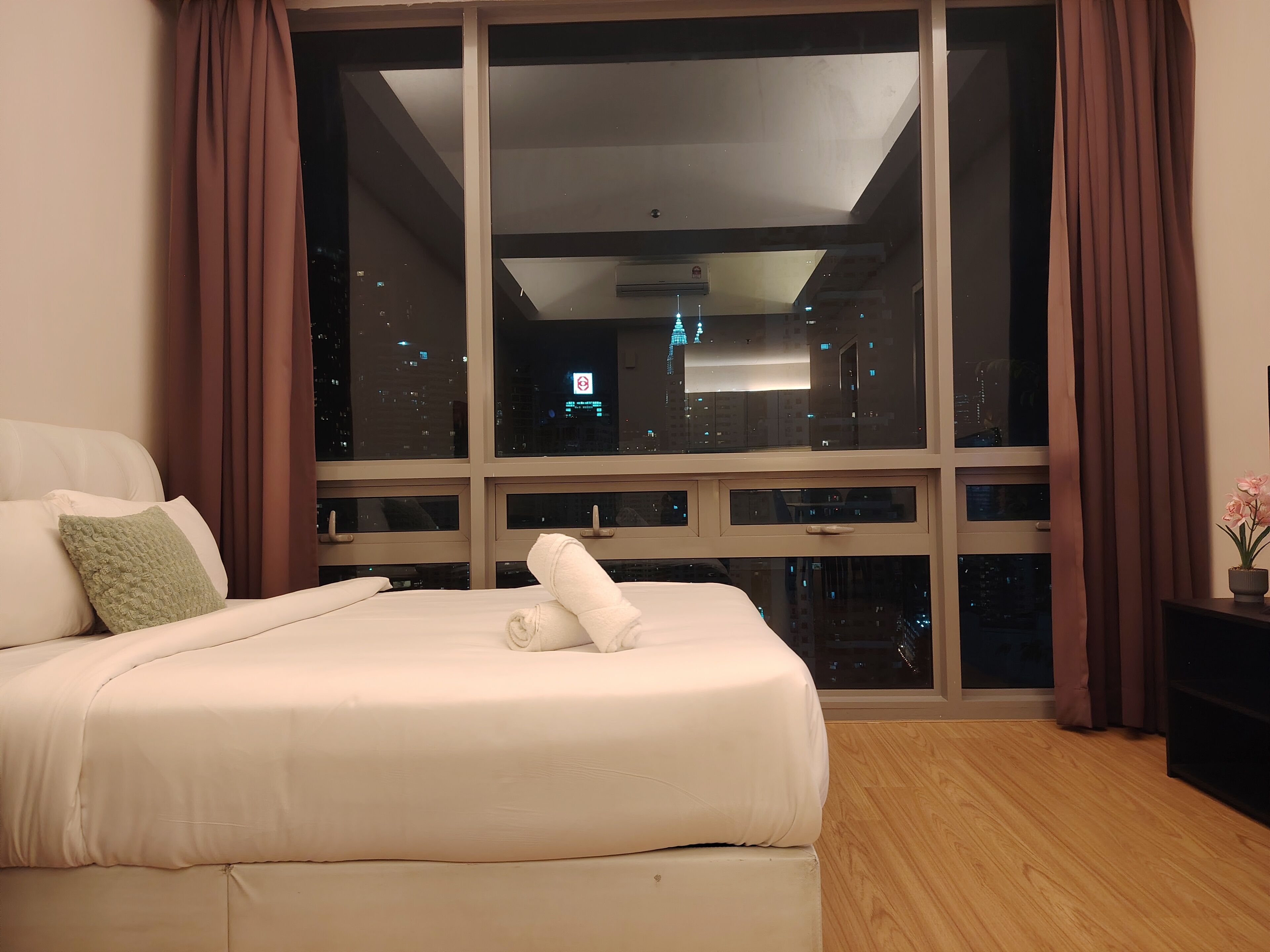 Ein Hotelzimmer mit großem Fenster, einem Bett mit weißen Leinen, einem grünen Kopfkissen und Blick auf die nächtliche Stadt.