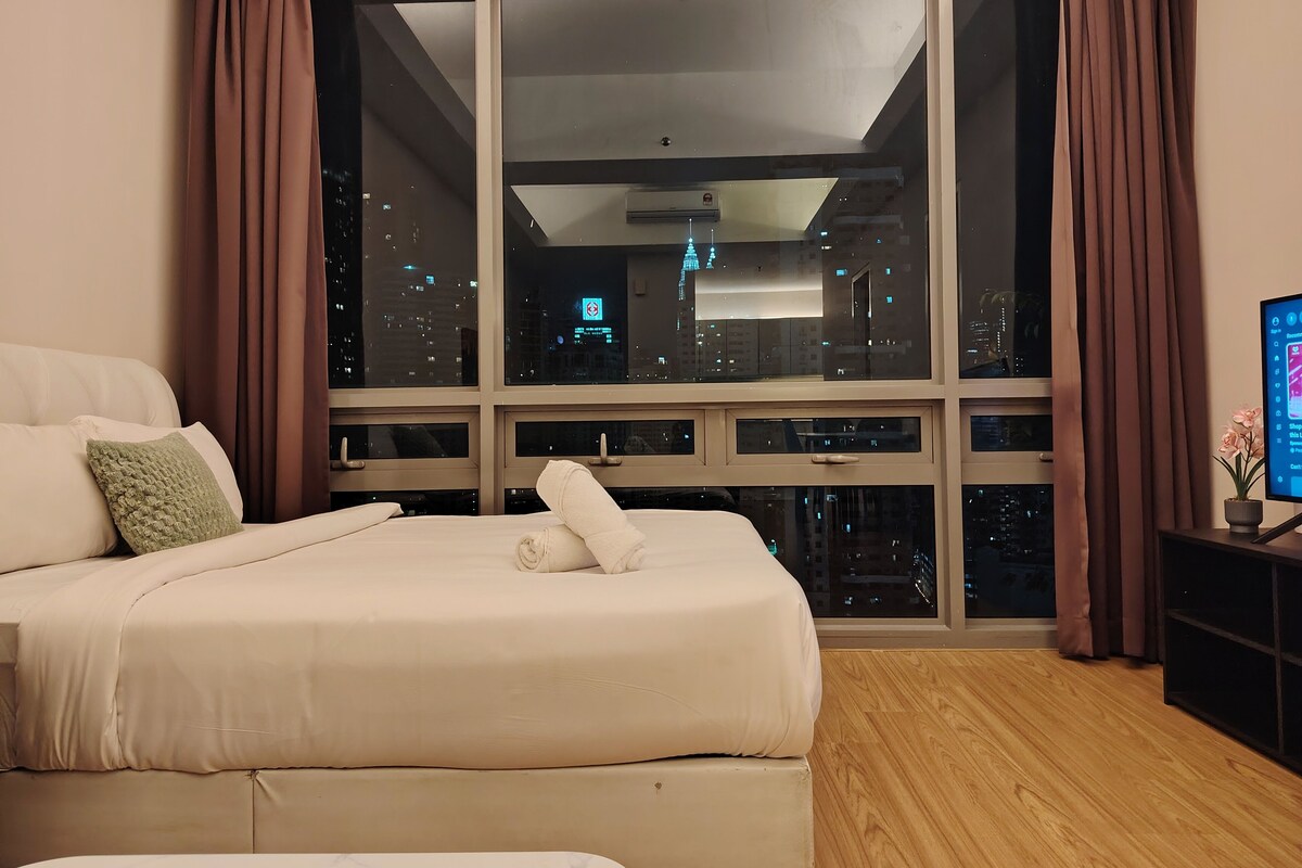A hotel room with a large bed, a view of a cityscape through floor-to-ceiling windows, and a television on a side table.