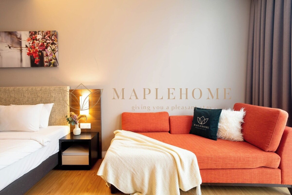 A hotel room with a bed, a nightstand with a lamp, a sofa, and a wall with the text 'MAPLEHOME'.
