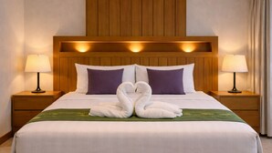 Una habitación de hotel con cabecera de madera, dos mesitas de noche con lámparas, cama con ropa de cama blanca y almohadas moradas, y una decoración de toallas con forma de cisne sobre la cama.