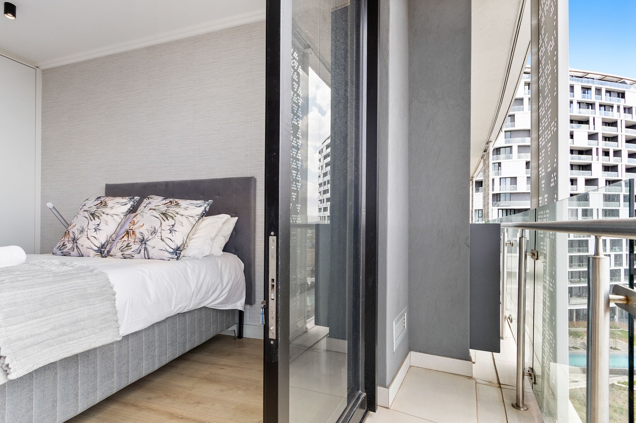 A modern bedroom with a bed, pillows, and a glass door leading to a balcony with a city view.