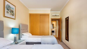 Een hotelkamer met twee bedden, een nachtkastje met een lamp, een houten garderobe en een ingelijst kunstwerk aan de muur.
