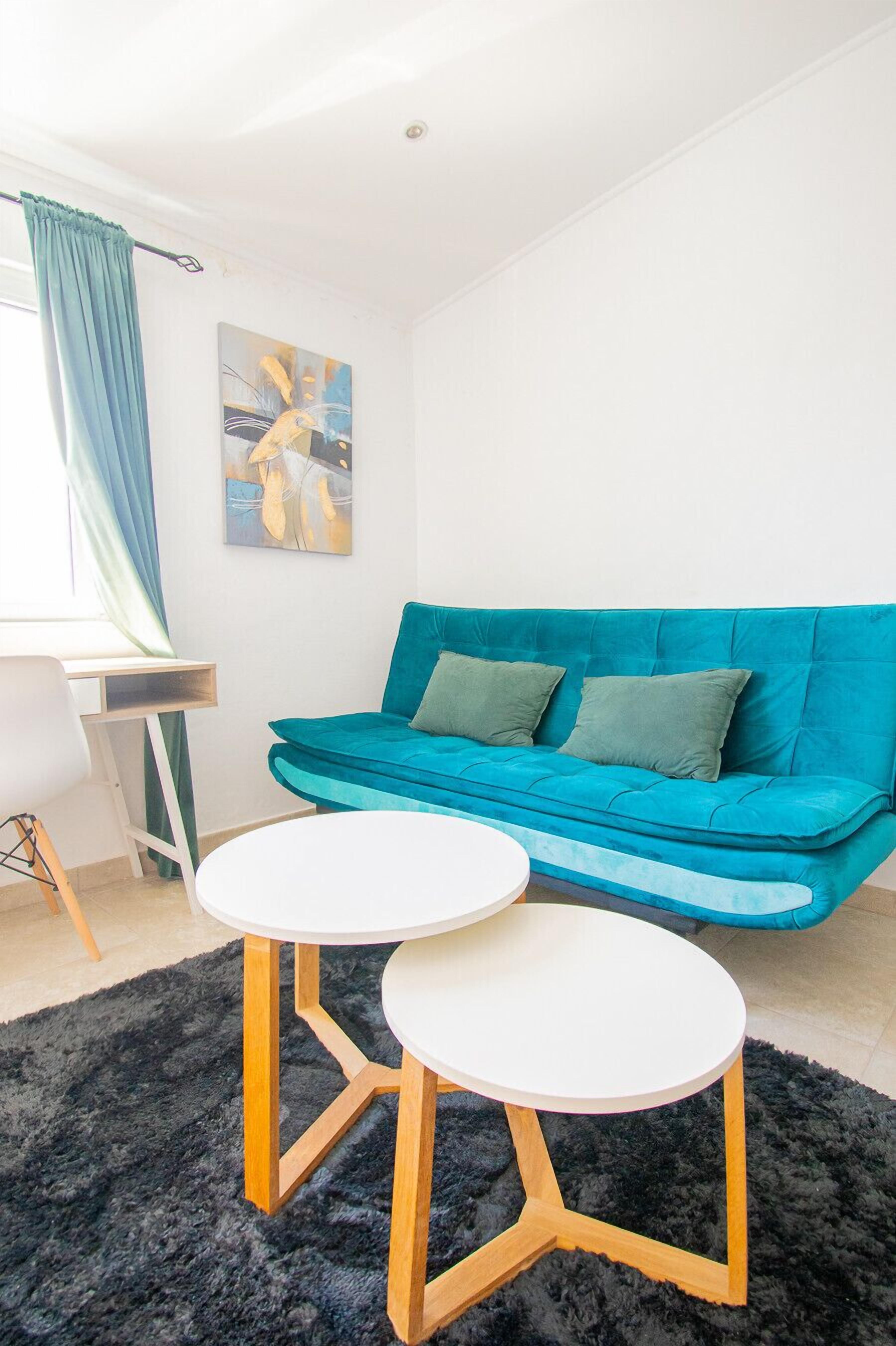Un salon moderne avec un canapé de couleur turquoise, deux petites tables rondes blanches, un tapis noir à poils longs et un tableau accroché au mur.