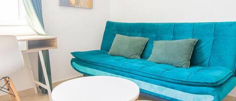 Un salon moderne avec un canapé de couleur turquoise, deux petites tables rondes blanches, un tapis noir à poils longs et un tableau accroché au mur.