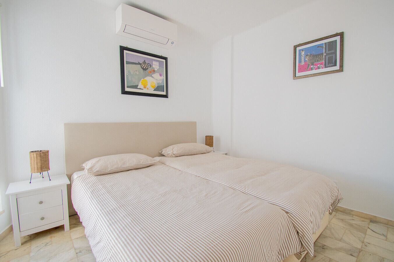 A bedroom with a bed, a nightstand, an air conditioner, and two framed pictures on the wall.