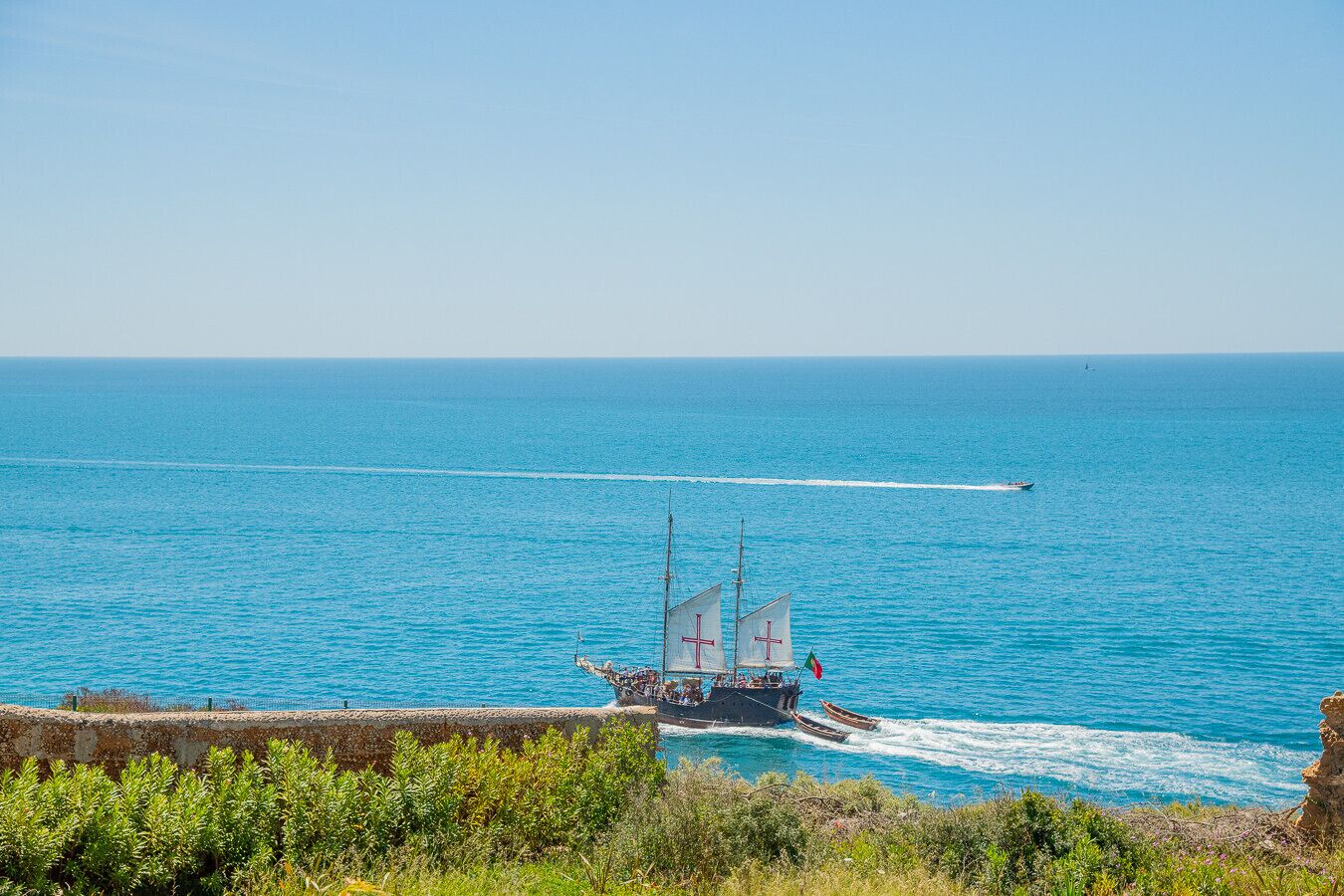 Paisaje costero con un barco navegando en el mar, cielo despejado y una costa rocosa con vegetación.