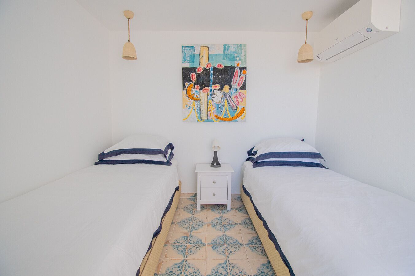 Une chambre avec deux lits, une petite table de chevet, une lampe et un tableau abstrait aux couleurs vives accroché au mur.