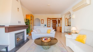 A living room with a fireplace, a white sofa, a glass coffee table, and a dining area with a lamp. - 115 Vale do Milho - Vista do Mar (Lagoa)