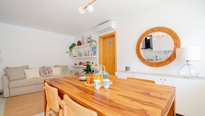 A modern living room with a wooden dining table, a sofa, and a kitchen area with a round mirror and a lamp. - Casa Flor do Mar in Carvoeiro (Lagoa)