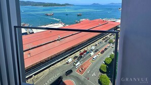 A view of a coastal area with a road leading to a building with a red roof, boats in the water, and a clear blue sky.