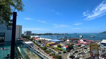 A view from a balcony overlooking a coastal town with a swimming pool, a busy street, and boats in the harbor.