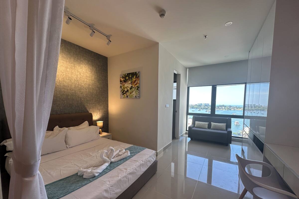 Habitación de hotel moderna con una cama grande, un rincón de estar con sofá y vistas al mar a través de amplios ventanales.