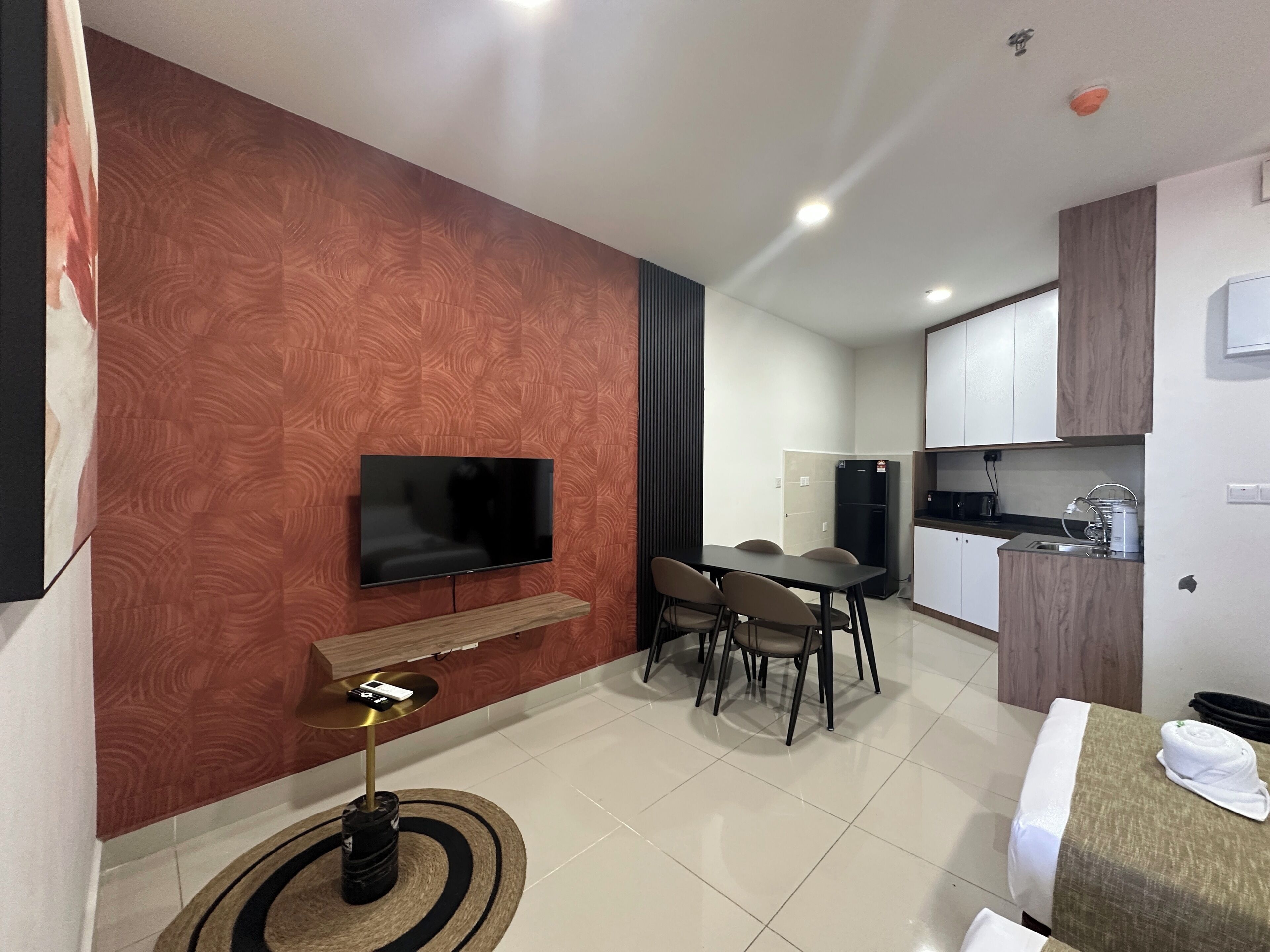 Ein modernes Hotelzimmer mit Flachbildfernseher an einer Holzwand, einem Esstisch mit vier Stühlen, einer kleinen Küchenzeile inklusive Kühlschrank und Kaffeemaschine sowie einem Bett mit weißem und gerolltem Handtuch.
