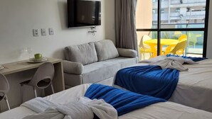 A hotel room with two beds, a desk with a chair, a TV mounted on the wall, and a view of the outside balcony. - Refuge By Paty - Malia Beach Resort for families (Ipojuca)