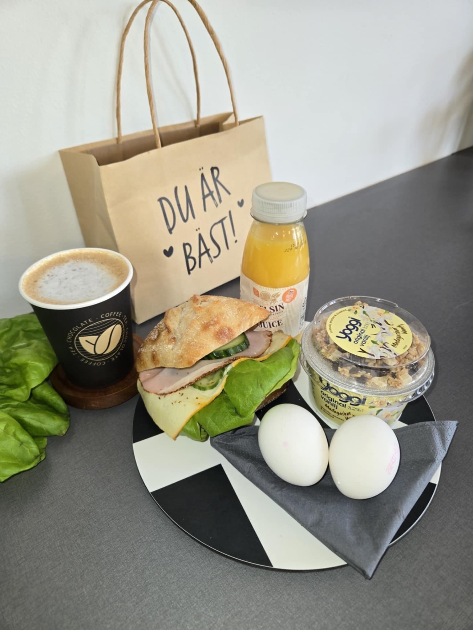 A meal with a sandwich, eggs, a drink, and a yogurt container on a plate.