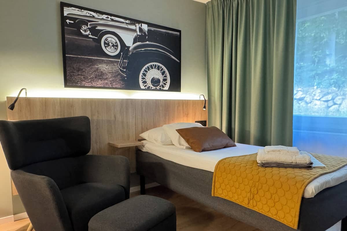 Une chambre d’hôtel comprenant un lit, une chaise, une petite table, une table de chevet, une lampe fixée au mur et une fenêtre avec des rideaux.