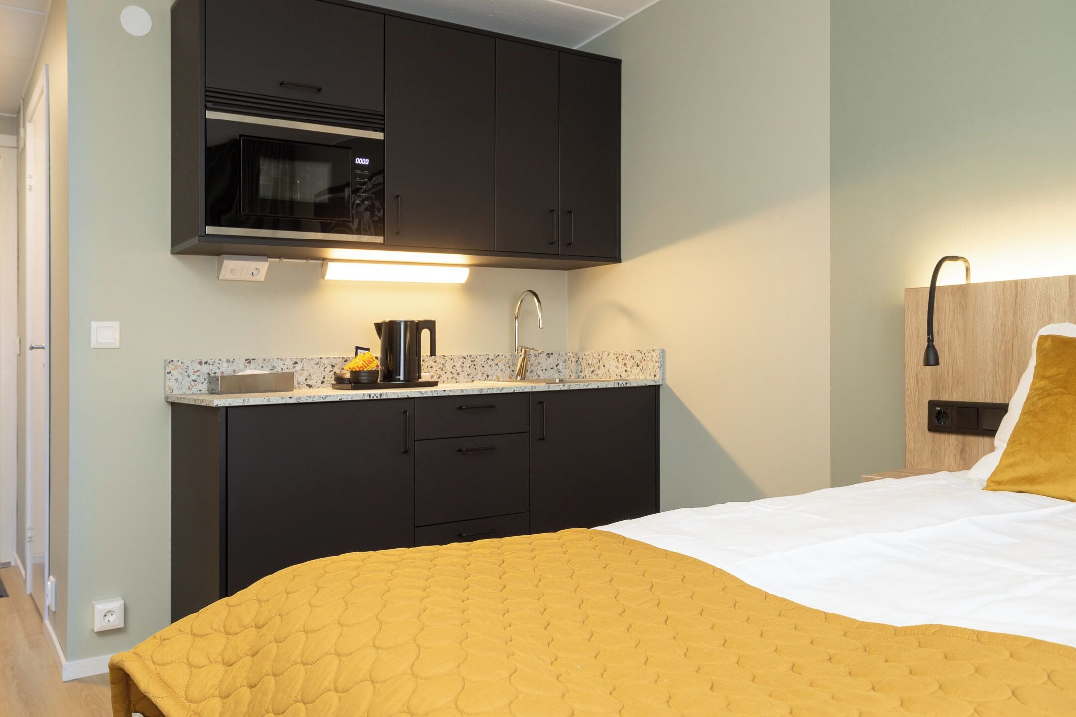 A compact hotel room with a bed, a kitchenette with a microwave, and a bedside table with a lamp.