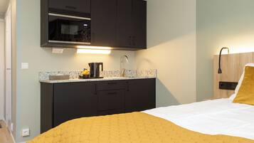 A compact hotel room with a bed, a kitchenette with a microwave, and a bedside table with a lamp.