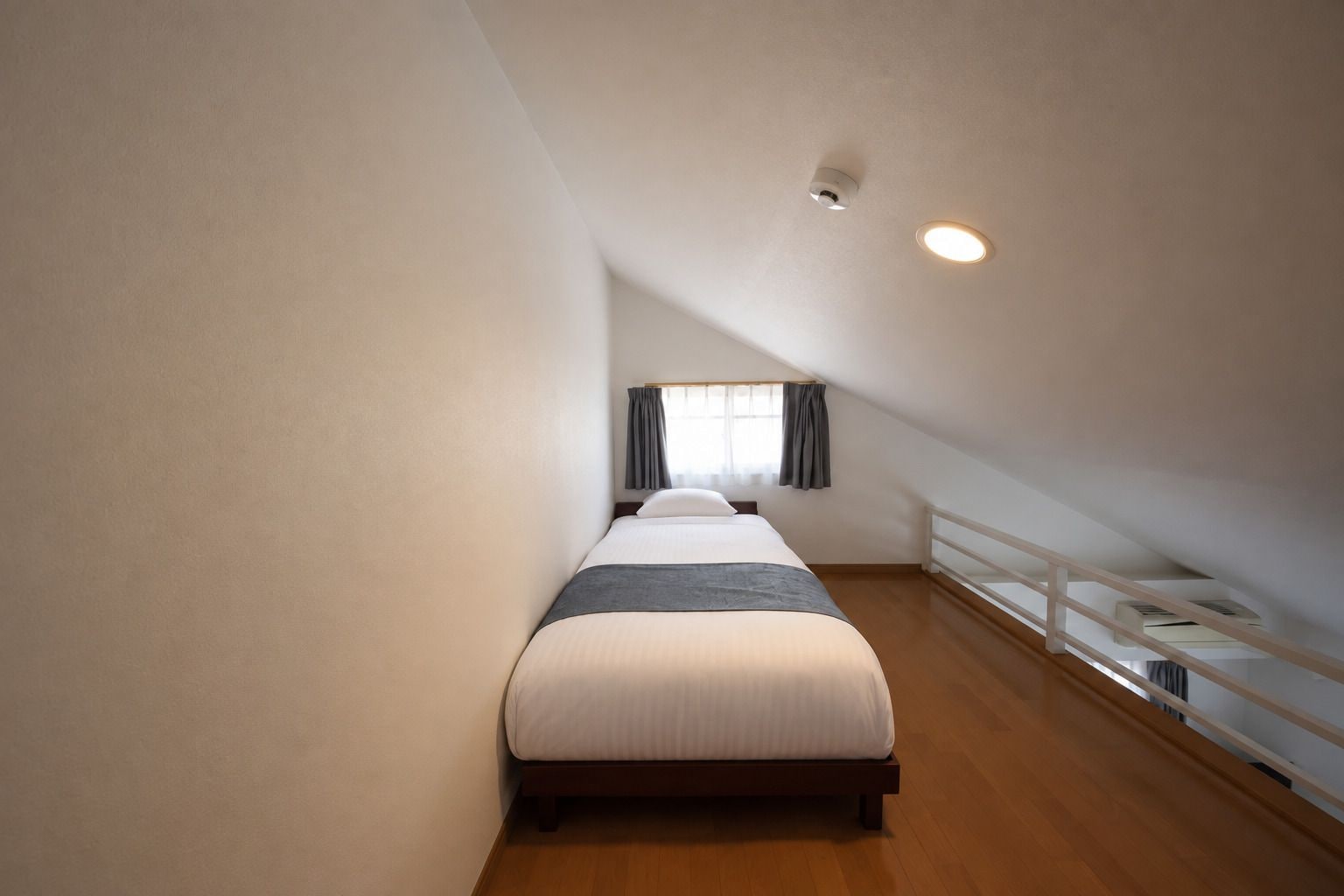 A narrow, single-bed room with a sloped ceiling and a window with curtains.