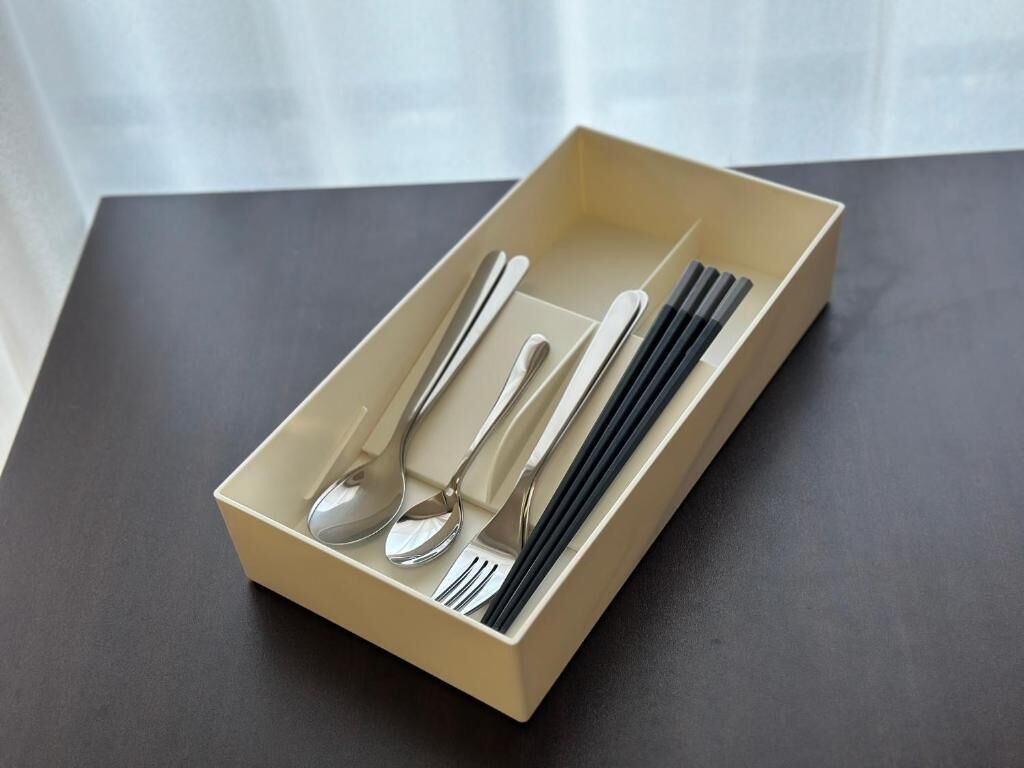 Una caja color crema con una cuchara, un tenedor y palillos sobre una superficie oscura.