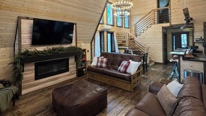 A spacious living room with a large television, a fireplace, a brown leather sofa, and a chandelier. - Cozy 2-bedroom secluded A-Frame cabin in charming Murray (Murray)