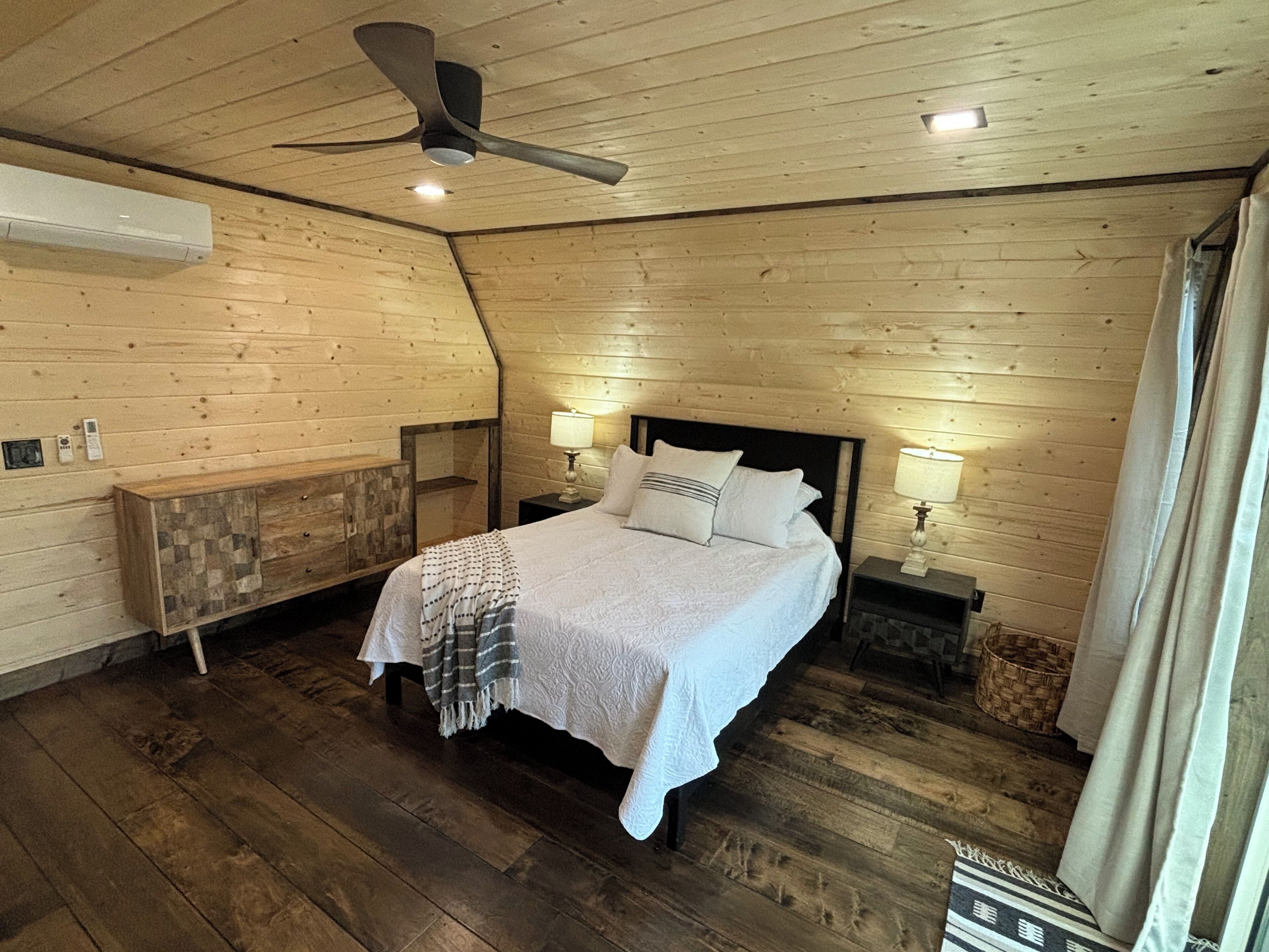 Een slaapkamer met een houten plafond, een bed met witte lakens, twee bedlampen en een plafondventilator.