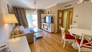 Ein modernes Wohnzimmer mit Sofa, Couchtisch und Esstisch mit Stühlen. Ein Fernseher ist vorhanden sowie eine kleine Küchenzeile mit Mikrowelle und Schrank.