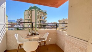 Ein Balkon mit Tisch, zwei Stühlen, einer Pflanze und Blick auf ein mehrgeschossiges Gebäude im Hintergrund.