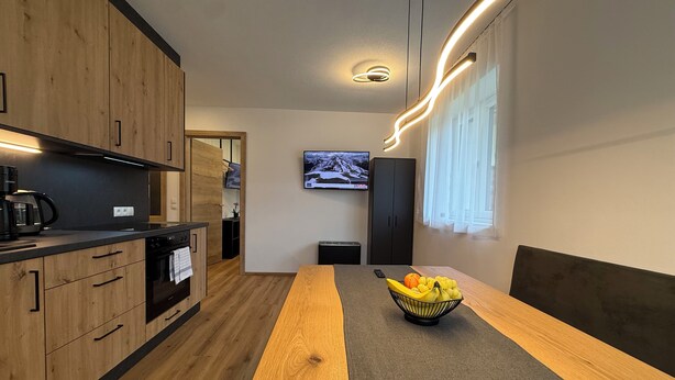 A modern kitchen with wooden cabinets, a built-in stove, and a dining area with a long wooden table and a fruit bowl. - Haus Schernthaner (Saalbach)