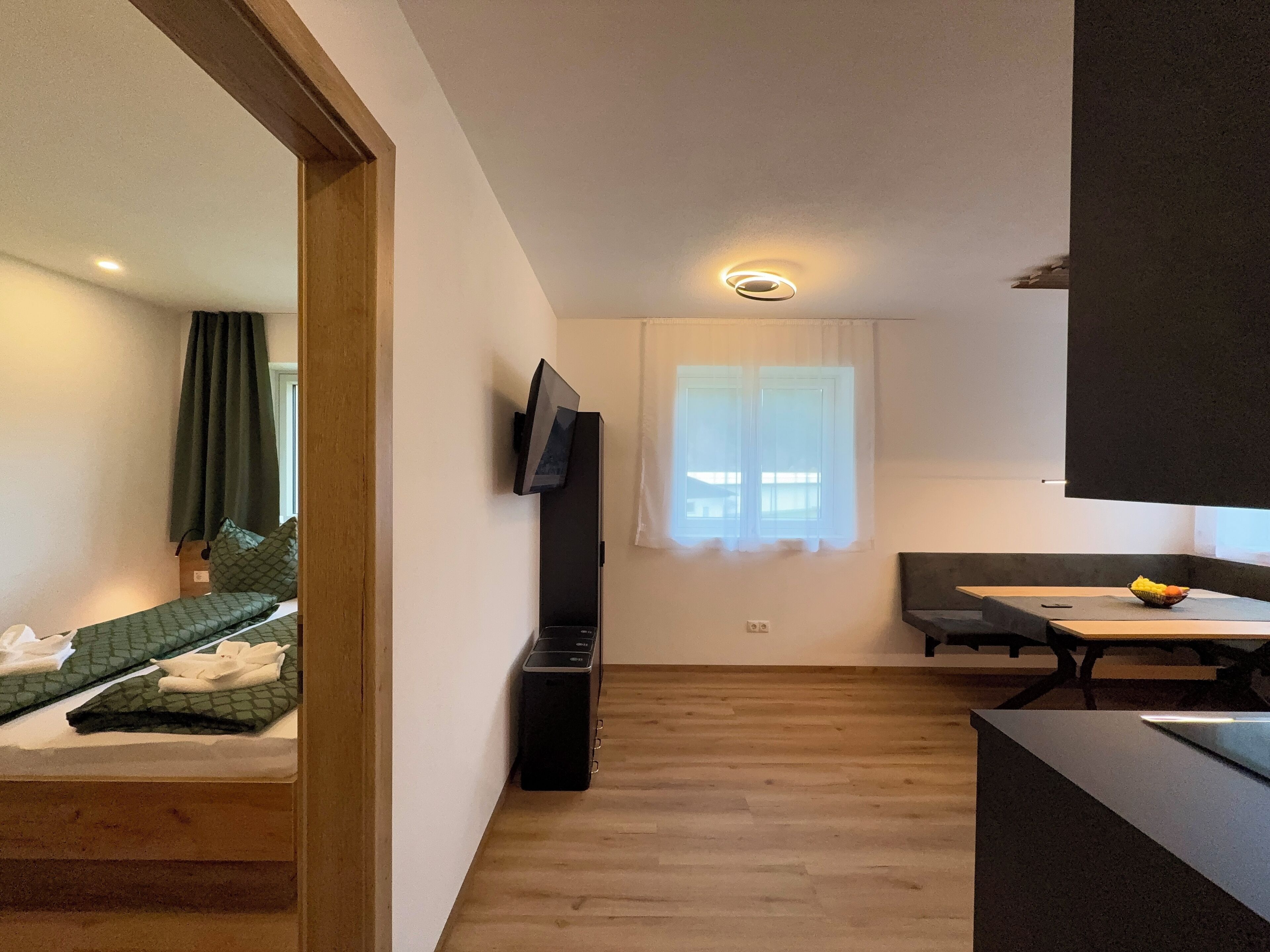 A compact living space with a bed, a small table, a TV mounted on the wall, and a window with curtains.