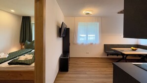 A compact living space with a bed, a small table, a TV mounted on the wall, and a window with curtains. - Haus Schernthaner (Saalbach)