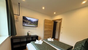 A hotel room with two beds, a TV mounted on the wall, a wooden door, and a small table with a vase and towels. - Haus Schernthaner (Saalbach)