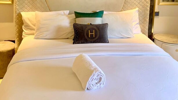 A neatly made bed with a quilted headboard, a rolled white towel, and a pillow with a 'H' emblem. - Sujimoto Residences - The AVEs  (Lagos)