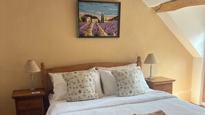 Een slaapkamer met een houten bed, wit beddengoed, een schilderij van lavendelvelden, twee nachtkastjes met lampen en een houten vloer.
