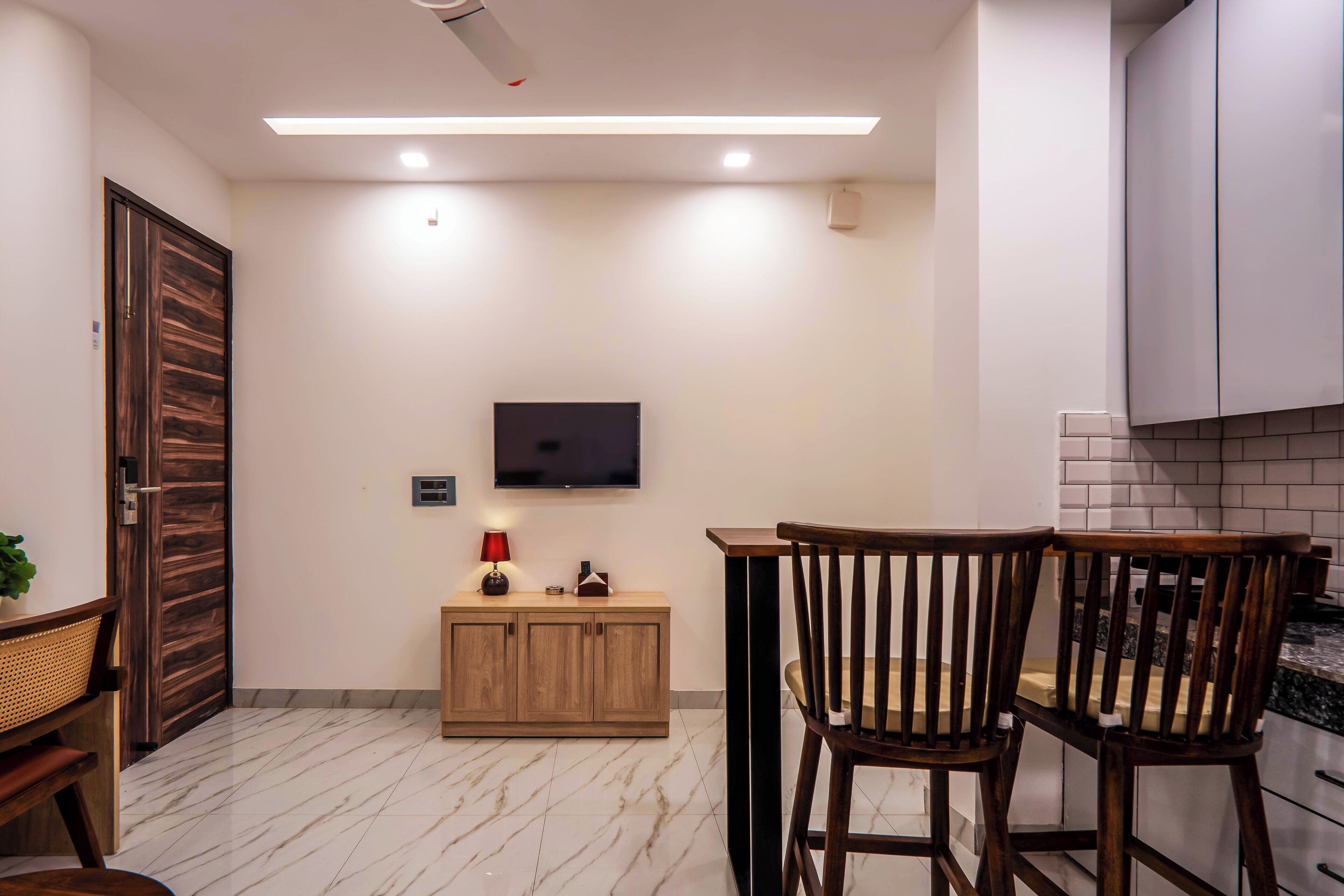 Una cocina moderna con un televisor montado en la pared, armarios de madera, suelo de mármol y un comedor con dos sillas de madera.