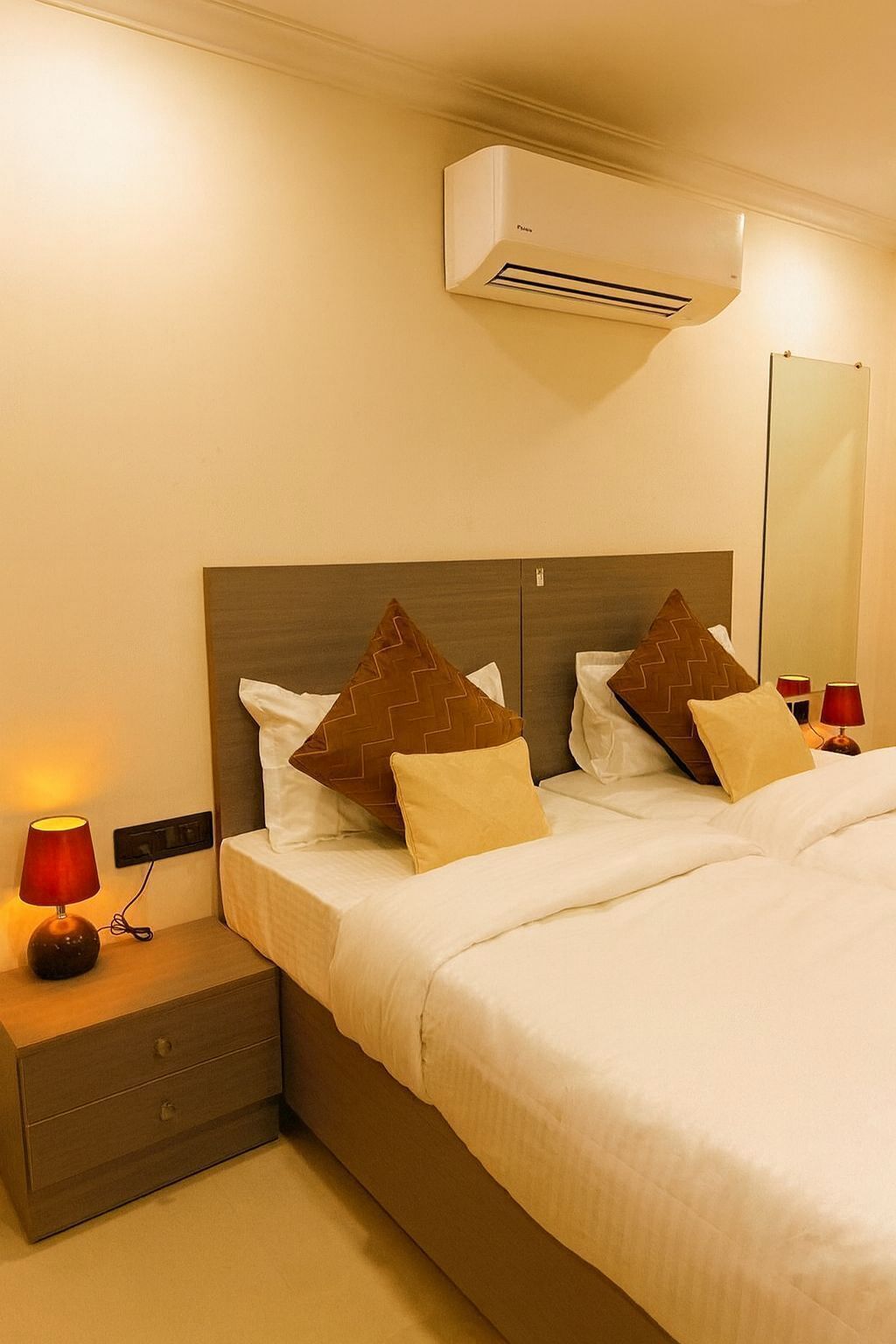 A hotel room with a bed, two bedside tables with lamps, an air conditioning unit, and a mirror on the wall.