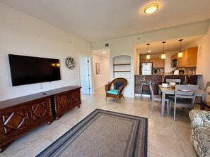 A living room with a flat-screen TV, wooden cabinet, and a blue armchair. - Stylish Beachside Condo with Balcony & Free round of Golf & Activities Daily! (Panama City Beach)