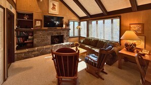 A cozy living room with a stone fireplace, wooden beams, a television, and a bookshelf. - Spacious Sugar Mountain Condo with Golf Course Views & Prime Location (Sugar Mountain)