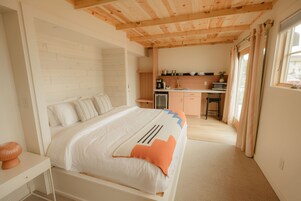 Une chambre à coucher comprenant un lit, une table de chevet, un plafond en bois, un coin cuisine et une fenêtre avec des rideaux.