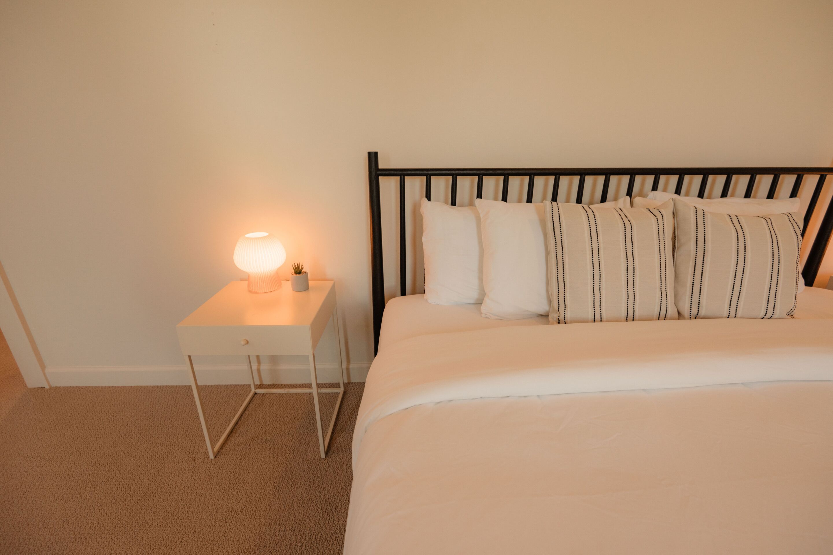 A neatly made bed with a black headboard, a bedside table with a lamp, and a small potted plant.