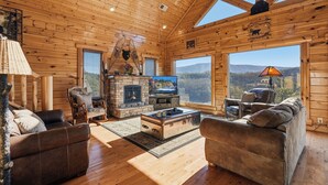 Ein Holzblockhaus-Interieur mit Kamin, Ledersofas und großen Fenstern mit Blick auf die Berge.
