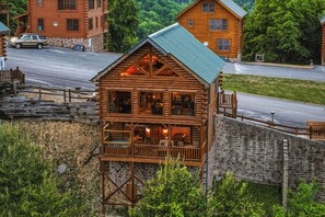 A log cabin with a balcony, a stone wall, and a road in the background. - New Listing! Breathtaking Mountain Views, Hot tub, Game Room, Jacuzzi Tub (Sevierville)