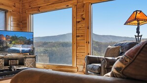 Ein gemütliches Blockhaus-Interieur mit Fernseher, Ledersofa und großem Fenster mit Blick auf die Berge.