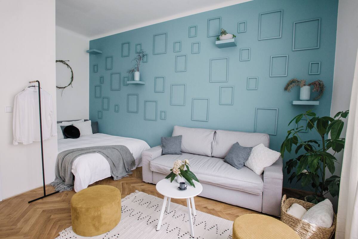 Un dormitorio moderno con una pared azul de acento, un sofá blanco, una cama con manta gris y una mesita redonda con un jarrón.