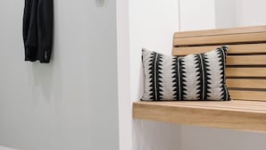 Een kamer met een houten bankje, gekleurde kussens, een hoed en een jas hangend aan de muur.