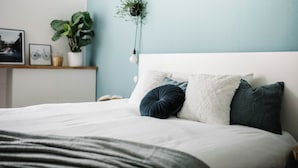 Een netjes opgemaakt bed met een wit hoofdbord, een donkerblauwe kussen en een witte kussen. Op een plank naast het bed staat een kleine plant, en aan de muur hangt een lijst met een foto.