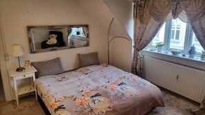 Et soveværelse med en seng, et natbord, et spejl, et vindue med gardiner og en radiator.