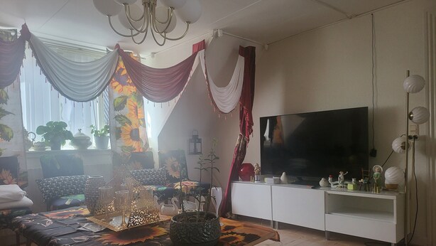 En stue med sofa, spisebord med blomstret bordløber, fjernsyn på hvidt møbel og en lysekrone.