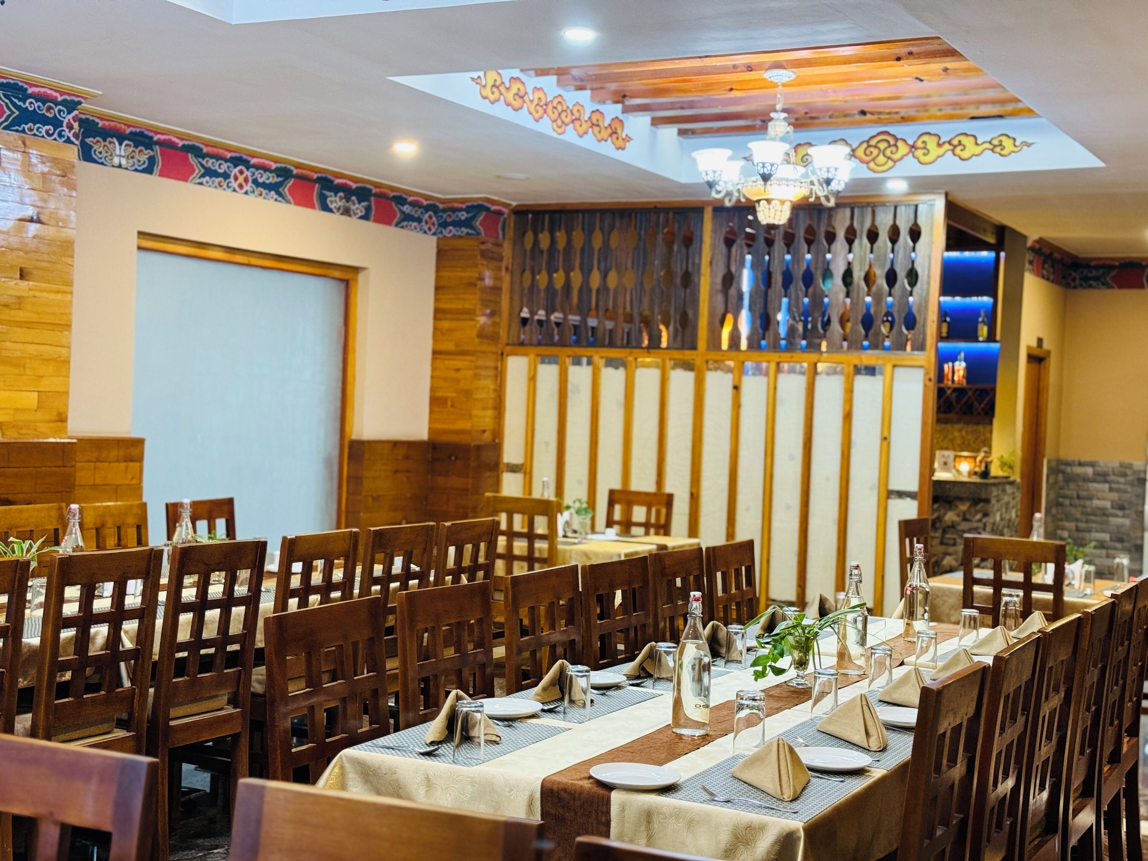 A restaurant with wooden furniture and a high ceiling with a chandelier. Tables are set with plates, glasses, and napkins.